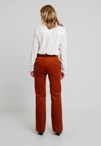 Orange Cord-Ripp-Hosen mit ausgestelltem Bein und strukturiertem Finish, kombiniert mit einem weißen gestreiften Hemd. Ansicht von hinten, die eine figurbetonte Taille und Taschen zeigt.