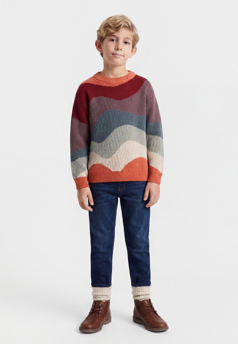 Garçon blond debout, portant un pull à motifs ondulés multicolores, un jean bleu, des guêtres crème et des bottes marron à lacets, sur un fond uni.