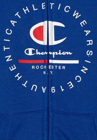 Sudadera azul con cremallera hecha de un tejido suave. Presenta un gran logo en blanco y rojo con las palabras "Champion" y "Rochester, N.Y." en el frente.