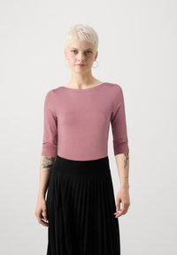 Piemērots, trīs ceturtdaļu piedurkņu tops bālganā rozā krāsā, izgatavots no mīksta auduma. Kombinēts ar melnu plisētu svārku ar plašu jostu.