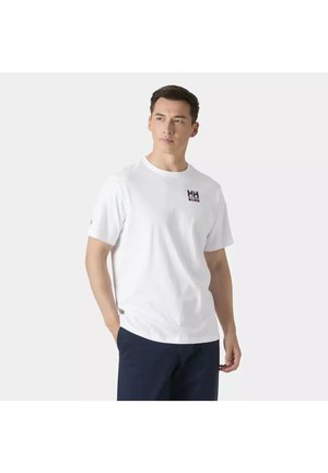 Hombre con camiseta blanca de manga corta con un pequeño logo en el pecho y pantalones oscuros, de pie con una mano en el bolsillo.