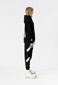 Sudadera negra de gran tamaño con impresión gráfica, combinada con pantalones de chándal negros que tienen bordado de alas blancas. Botas blancas de suela gruesa con cordones.