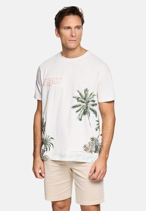 Hombre con camiseta blanca de manga corta con estampado de palmeras y pantalones cortos beige, de pie sobre un fondo claro liso.