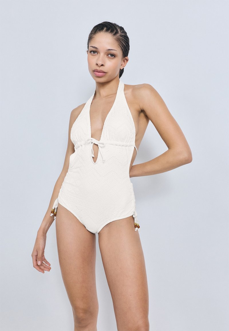 Λευκό ολόσωμο μαγιό με βαθύ V ντεκολτέ, κλείσιμο με δέσιμο, υφασμάτινο μοτίβο και διακοσμητικές χάντρες στα πλευρικά κορδόνια. Λείο ύφασμα.