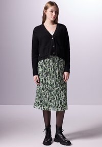 Černý žebrovaný cardigan se třemi knoflíky, spárovaný s zelenou plisovanou sukní se vzorem černé a zelené. Modelka má na sobě černé šněrovací boty.
