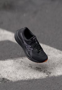 Baskets de sport noires avec tige en mesh, accents violets, surface texturée et semelle en caoutchouc. Comprend des lacets et une forme épurée.