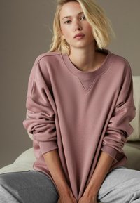 Przyduży różowy sweter z okrągłym dekoltem, prążkowanymi mankietami i luźnym krojem, zestawiony z szarymi dresami na beżowej sofie.