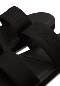Schwarze Slide-Sandalen aus strukturiertem Stoff mit zwei breiten Riemen. Das Fußbett ist flach und sanft konturiert, mit dezenten Markenakzenten.