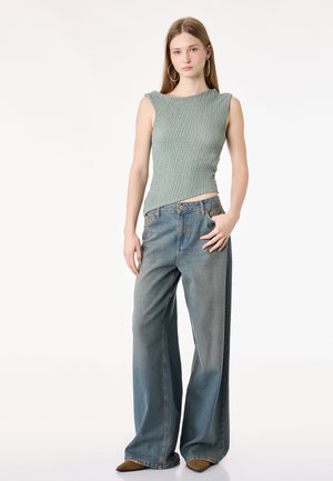 Junge Frau, die ein hellgrünes, strukturiertes ärmelloses Oberteil, weit geschnittene, verwaschene blaue Jeans, braune spitze Schuhe und große Creolen trägt.