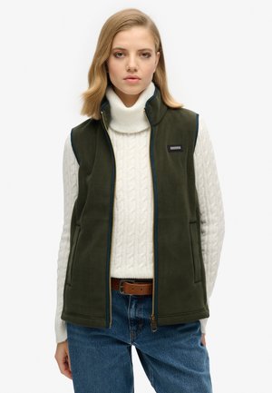 Chaleco de forro polar verde oliva con cuello alto, cierre de cremallera por delante y ribete en azul marino; llevado sobre un suéter de punto trenzado blanco y jeans azules.