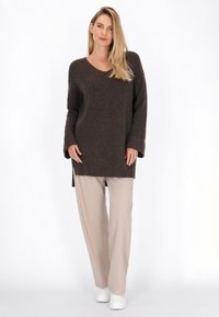 Pulover tricotat maro cu decolteu în V și mâneci lungi, purtat cu pantaloni bej deschis și adidași albi. Dispune de schituri laterale pentru un detaliu suplimentar.