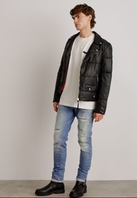 Svart lærpufferjakke med glidelås og lommer, båret over en hvit skjorte og lyseblå slitte jeans, komplementert med svarte støvler.