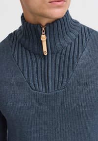 Dunkelblauer Strickpullover mit hohem Kragen, vertikaler Rippung und halbem Reißverschluss mit einem beigen Lederzieher.