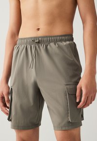 Shorts cargo en vert olive discret, avec une ceinture élastique, un cordon de serrage et des poches latérales pour le rangement. Tissu lisse et léger.