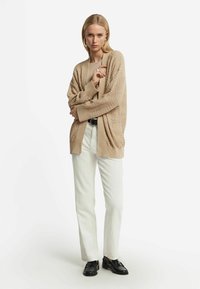 Beige stickad cardigan med en texturerad design, öppen fram, och långa ärmar, tillsammans med en beige topp, vita byxor och svarta skor.