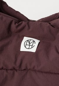 Bordeauxfarbener Pufferstoff mit einem auffälligen weißen Etikett, das ein schwarzes Logo zeigt. Die Textur ist glatt, mit sichtbaren Nähte Details.