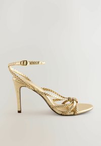 Guld högklackad sandal med flätade remmar, ankelspråk och spetsig tå. Slät metallisk finish med en smal stilettklack.