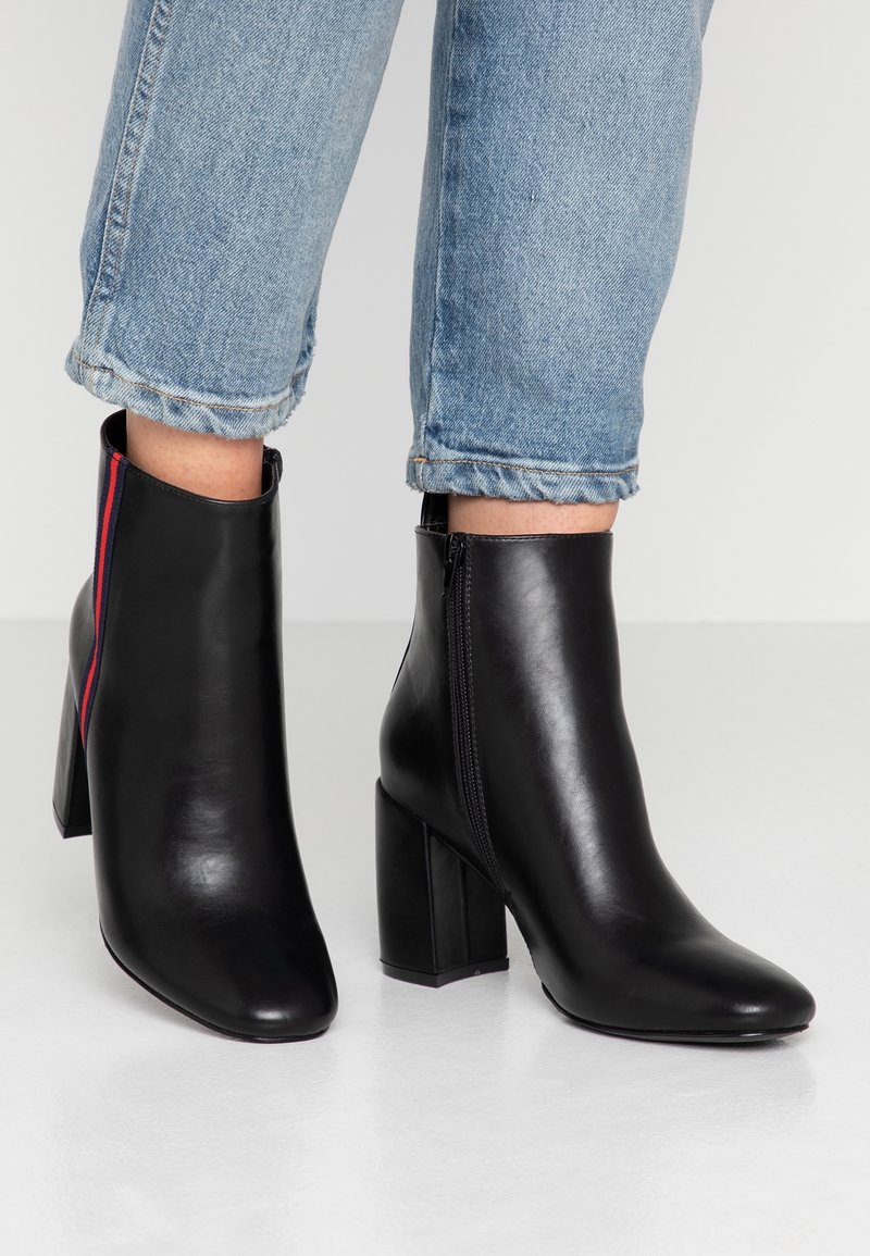 Bottines en cuir noir avec talons carrés, fermeture éclair latérale, et détail à rayures rouge et bleu, portées avec un jean bleu clair retroussé.