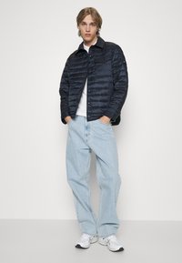Giubbotto imbottito navy con colletto strutturato, abbinato a jeans larghi azzurri e sneakers bianche. Il giubbotto presenta una texture trapuntata.