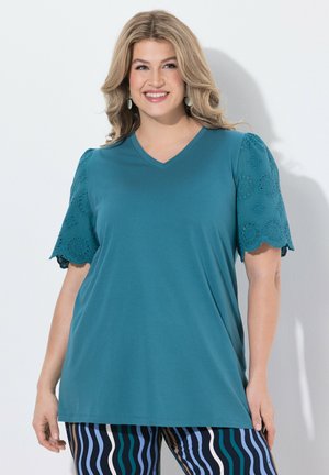 Femme souriante aux cheveux blonds ondulés portant un haut vert sarcelle à col en V avec des manches en dentelle à œillets et un pantalon rayé bleu, noir et blanc sur fond blanc.