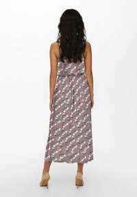 Donna con lunghi capelli scuri, che indossa un vestito midi senza maniche color malva con fiori e tacchi alti beige, in piedi di spalle su uno sfondo bianco.