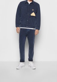 Navy blue farkkutakki, jossa on nappisuljenta, rintatasku ja vastaavat farkut, yhdistettynä valkoisiin korkeisiin lenkkareihin.