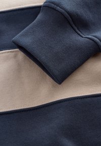Foldet manchet på en mørkeblå og beige color-blocked sweatshirt, der viser detaljeret stofstruktur og syning.