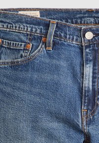 Jean en denim bleu en gros plan montrant la poche avant, la boucle de ceinture, le bouton en métal, les rivets et l'étiquette sur la ceinture.