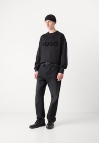 Black sweatshirt with bold "HUGO" logo, paired with dark denim jeans. Model wears a black knit beanie and black shoes. Simple, modern look.