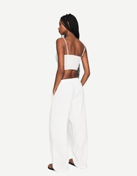 Une jeune femme aux longues tresses se tient de dos, vêtue d'un top court blanc et d'un pantalon blanc ample, sur un fond blanc uni.