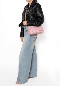 Borsa a tracolla rosa in pelle con superficie texturizzata, forma rettangolare e patta frontale. Il modello indossa una giacca di pelle nera e jeans a gamba larga.