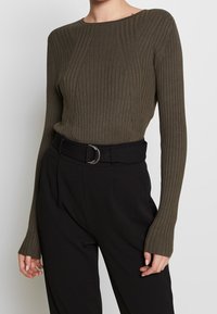 Długie rękawy prążkowanego, oliwkowozielonego topu z okrągłym dekoltem, zestawione z czarnymi, wysokim stanem spodniami z paskiem zakończonym okrągłą klamrą.