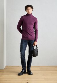 Pull à col roulé en laine mérinos de couleur violet foncé, au design ajusté. Associé à un jean foncé et des chaussures noires. Comprend un petit sac bandoulière noir comme accessoire.