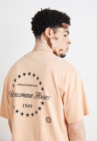 T-shirt de algodão na cor pêssego com texto preto e design de estrela nas costas, incluindo "ADIDAS BASKETBALL" e "WORLDWIDE HOOPS 1949."