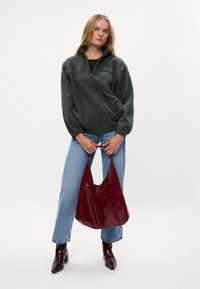 Groene fleece vest met rits en lange mouwen, gecombineerd met lichtblauwe spijkerbroek en een glanzende bordeauxrode schoudertas. Zwarte enkelboots maken de look compleet.