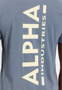 Szürke pamut póló, amelyen egy nagy, függőleges krémszínű logó található, amely a hátán az "ALPHA INDUSTRIES" feliratot viseli. Rövid ujjú, normál fazonú.