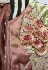 Gros plan d'une veste en satin rose avec des roses en tissu effiloché brodées et des feuilles vertes près de la fermeture éclair argentée et du col rayé noir et blanc.
