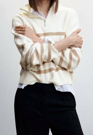 Personne les bras croisés portant un pull blanc à rayures beige sur une chemise blanche et un pantalon noir.