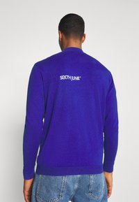 Lila långärmad sweatshirt med ribbade ärmslut och nederkant, med en vit logotyp "SIXTHJUNE" tryckt på baksidan. Mjuk textur, avslappnad passform.