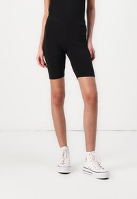 Calças de ciclismo pretas feitas de tecido elástico, com uma cintura alta. Combinadas com ténis brancos de cano alto e uma blusa preta ajustada.