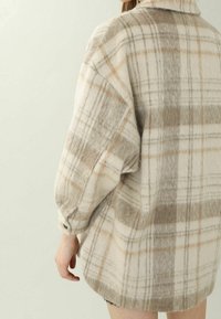 Brushed flannel shirt in cream with beige and gray plaid pattern; oversized fit, dropped shoulders, button cuffs, and curved hem.