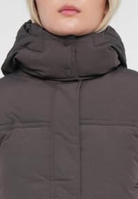 Doudoune gris foncé avec un col haut, présentant des coutures visibles, des boutons-pression et une texture de tissu lisse et légèrement brillante.