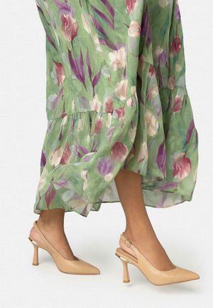 Piernas con tacones puntiagudos color canela con tiras y hebillas, vistiendo un vestido midi verde floral y fluido en movimiento.