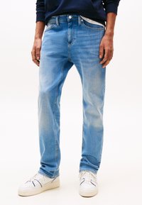 Homme portant un jean bleu clair délavé, des baskets blanches et un sweat-shirt bleu foncé, les mains détendues le long du corps.