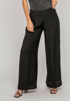 Femme portant un pantalon noir taille haute à jambes larges et des sandales noires à lanières, posant avec une main tenant la ceinture.