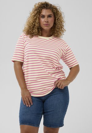 Femme aux cheveux blonds bouclés portant un t-shirt crème à manches courtes avec des rayures roses et un short en jean jusqu'aux genoux, se tenant avec une main sur la hanche.