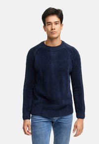 Granatowy sweter z dzianiny o strukturalnym wzorze, okrągłym dekolcie i ściągaczami przy mankietach. Noszony z jasnoniebieskimi jeansami, prezentującymi luźny krój.
