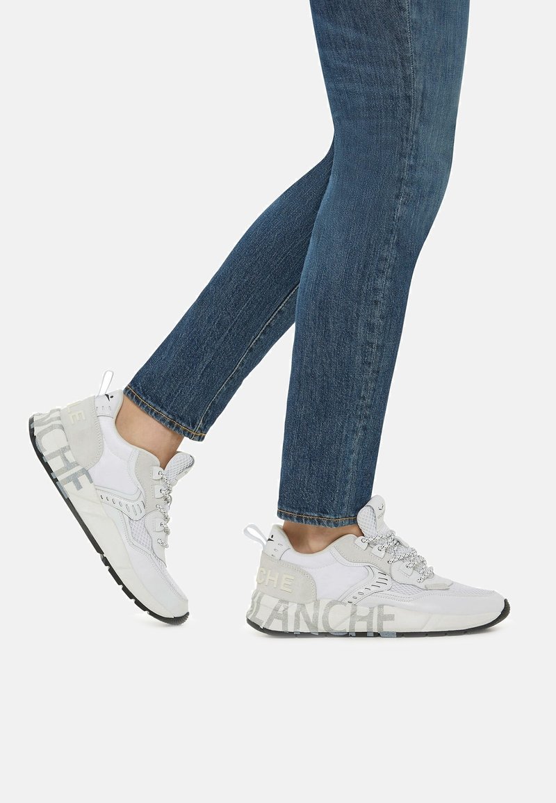 Personne portant un jean bleu et des baskets blanches avec des semelles noires et un texte gris "BLANCHE" sur le côté, montrée en train de marcher sur un fond uni.