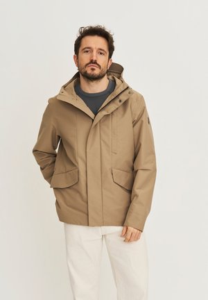 Man wearing tan hooded jacket over dark sweater and white pants, standing with one hand in pocket against plain background.