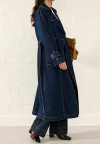 Mujer que lleva un largo abrigo de mezclilla oscuro sobre unos pantalones anchos a juego y botas de punta cuadrada oscuras, sosteniendo un bolso de cuero color beige.